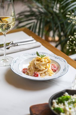 Parlak bir restoran masasında beyaz bir tabakta servis edilen vişneli domatesli kremalı makarna ve taze otlar.