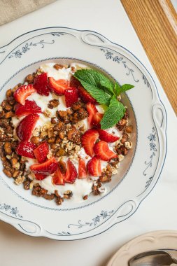 Sağlıklı granola kasesi kremalı yoğurt taze çilek ve fındıklar parlak bir masada dekoratif bir tabakta servis edilir.
