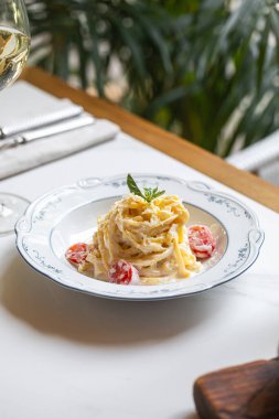 Parlak bir restoran masasında beyaz bir tabakta servis edilen vişneli domatesli kremalı makarna ve taze otlar.
