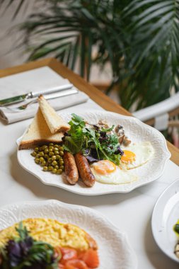Kızarmış yumurtalı kahvaltı tabağı sosis, bezelye tostu ve beyaz tabakta servis edilen taze salata.