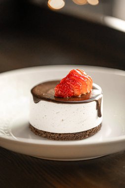 Kremalı çikolatalı mini kek, parlak çikolata jöleli ve beyaz bir tabakta servis edilen taze çilekli.