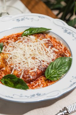 Klasik lazanya, bol domates soslu rendelenmiş peynir ve seramik tabakta sunulan taze fesleğen ve dramatik yan ışık ve derin gölgelerle servis edilir.