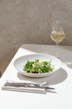 Taze yeşil salata, karışık otlar ve hafif sosla birlikte restoran masasında bir bardak beyaz şarapla servis ediliyor.