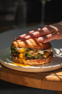 Erimiş peynir ve soslu sulu çizburger gün ışığında tabakta elle eziliyor.