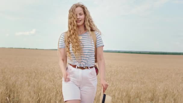 Чарівна красива дама з великою посмішкою і довгим кучерявим волоссям насолоджується часом, коли вона проходить через пшеничне поле — стокове відео
