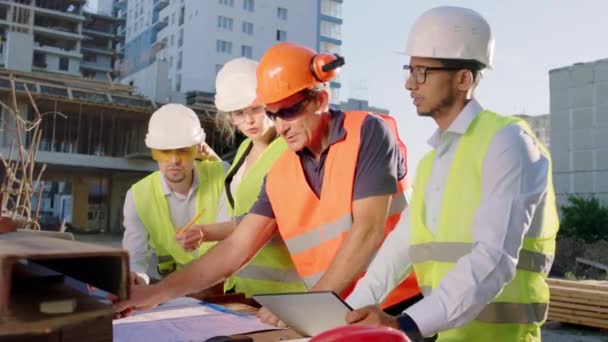 Vor der Kameragruppe diverser Spezialisten auf der Baustelle analysieren sie gemeinsam einen Bauplan und tragen dabei Schutzhelme und Schutzbrillen. — Stockvideo