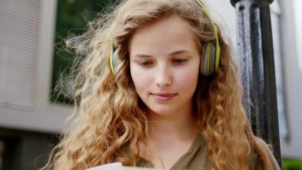 Харизматичная с красивым лицом молодая женщина, использующая наушники для прослушивания музыки она улыбается большой, печатая что-то на портрете смартфона перед камерой. Снимок на ARRI Alexa Mini — стоковое видео