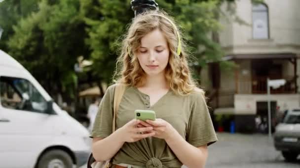 Гарно виглядає блондинка перед камерою посеред головної вулиці щось на смартфоні і посміхається, що вона носить великий жовтий навушники. — стокове відео