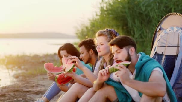 Al atardecer junto al grupo de amigos multiétnicos del lago felices comiendo un trozo de sandía fresca disfrutando del tiempo en el picnic de la naturaleza — Vídeo de stock