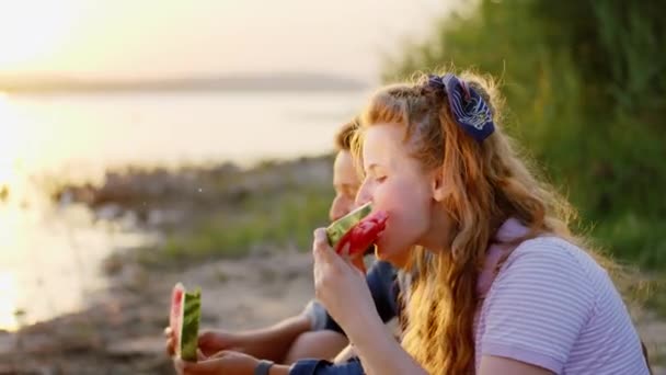 O zachodzie słońca przy pikniku niesamowicie wyglądająca pani i jej przyjaciel Afro Amerykanin jedzą arbuza i patrzą na zachód słońca — Wideo stockowe