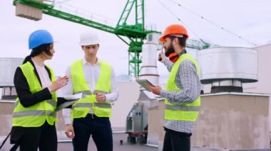 Genç mühendis ve mimarlardan oluşan iyi görünümlü bir grup binanın tepesindeki inşaat alanının planlarını inceliyor. İnşaat alanında birlikte yürüyorlar.