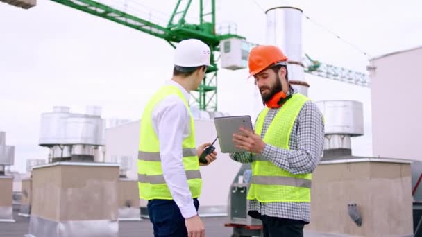 Modern construction site one builder and businessman on the roof top using digital tablet to analyzing the plan of construction they happy smiling and discussing the businessman take a radio — Stock Video
