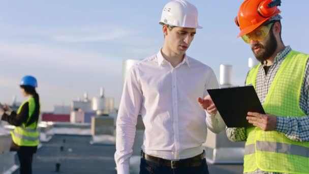 In front of the camera in the top of the construction building businessman have a discussing with the engineer about the site his assistant pretty lady take the plan of construction from the digital — Stock Video