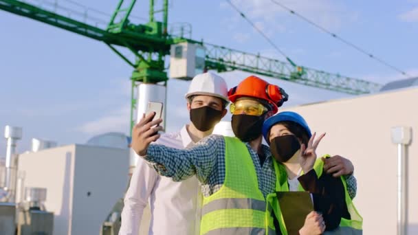 Gruppe junger verschiedener Spezialisten auf der Baustelle am Ende des Tages in der Pandemie des Coronavirus machen einige Selfies, die sie mit schwarzer Schutzmaske und Sicherheitsausrüstung machen — Stockvideo