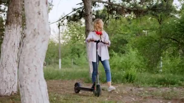 Sehr aufgeregte Blondine kam mit ihrem Elektroroller in den Park und genoss die Zeit, in der sie rote Kopfhörer trug — Stockvideo