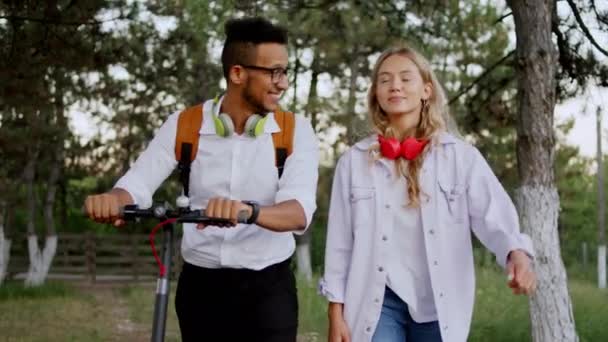 Parkta iki arkadaş, kadın ve erkek, birlikte yürüyorlar ve kameranın önünde dostça sohbet ediyorlar. Afrikalı Amerikalı adam, elektrikli scooterını tutuyor ve mutlu bir ruh halindeler. 4k — Stok video
