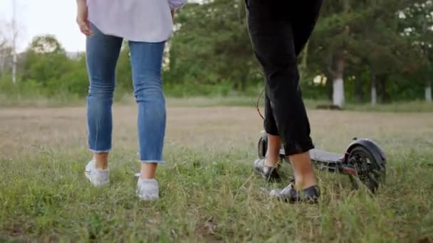 Подробиці двоє студентів, які ходять після курсу коледжу, ходять по парку, вони мають дружнього розмовника, який тримає свій електричний скутер — стокове відео