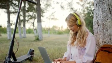 Parkta gülümseyen güzel bir bayan öğrenci battaniyenin üzerine oturur ve kulaklıktan müzik dinlerken dizüstü bilgisayarda bir şeyler yazar.