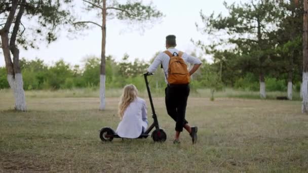 Dos estudiantes en el parque disfrutando del aire fresco en la naturaleza hablando entre sí señora tomar un asiento en el scooter eléctrico — Vídeos de Stock