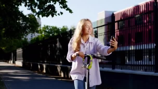 Ξανθά μαλλιά γυναίκα στη μέση του δρόμου κάνει μια στάση με ηλεκτρικό σκούτερ της για να κάνει μερικές φωτογραφίες με το smartphone που χαμογελά μεγάλο και αισθάνεται πολύ χαρούμενος — Αρχείο Βίντεο