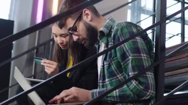 穿上衣服两个迷人的年轻人和一个日本女人坐在一个现代化的楼梯上，用信用卡从笔记本电脑上查看网上账户 — 图库视频影像