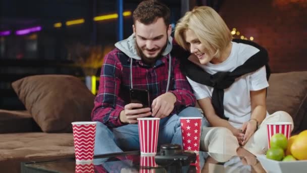En una moderna sala de estar loft en el sofá atractiva pareja mirando a través del teléfono inteligente y pasar un buen rato juntos — Vídeos de Stock
