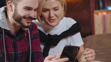 Kameranın önünde iki arkadaş, çekici sarışın kadın ve yakışıklı bir erkek birlikte kanepede oturup akıllı telefondan bakıp büyük bir gülümseme ile vakit geçiriyorlar.