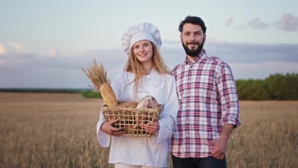 Belle belle dame boulanger et jeune homme agriculteur au milieu du champ de blé debout devant la dame caméra tenant un panier avec du pain frais. 4k — Video