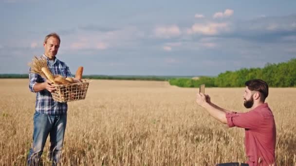 Mutlu ve heyecanlı çiftçi kameranın önünde poz veriyor oğlu elektronik tabletten fotoğraf çekiyor çiftçi buğday tarlasının ortasında bir kutu ekmek tutuyor. — Stok video