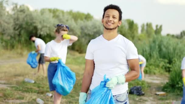 Przed kamerą uśmiecha się duży Afro amerykański facet trzymając niebieską plastikową torbę zbiera śmieci z plaży z innymi ochotnikami — Wideo stockowe