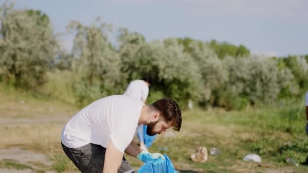 Le concept de recyclage groupe de bénévoles divers nettoyer ensemble une grande plage verte au bord du lac ils ramassent les ordures et de recueillir sur les sacs en plastique bleu. Tourné sur ARRI Alexa Mini — Video