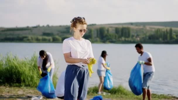 Al lado de un gran lago en el lado de la playa rubia pelo voluntario dama usando sus guantes protectores amarillos para empezar a limpiar la basura de la playa otro grupo de voluntarios usando grandes bolsas de plástico — Vídeo de stock