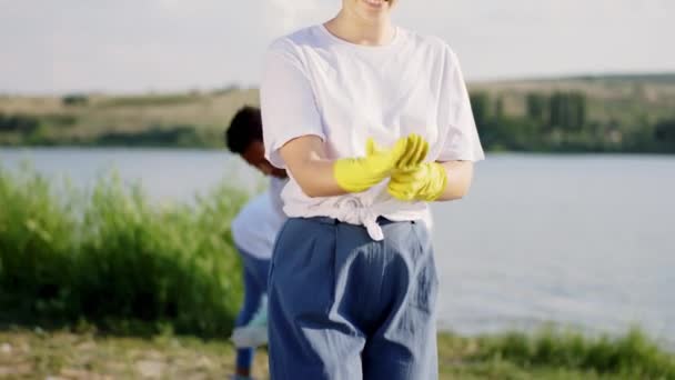 Delante de la cámara sonriendo gran dama linda con guantes protectores amarillos y empezar a recoger la basura del lado de la playa y recoger en las bolsas de plástico azul. Disparo en ARRI Alexa Mini — Vídeos de Stock