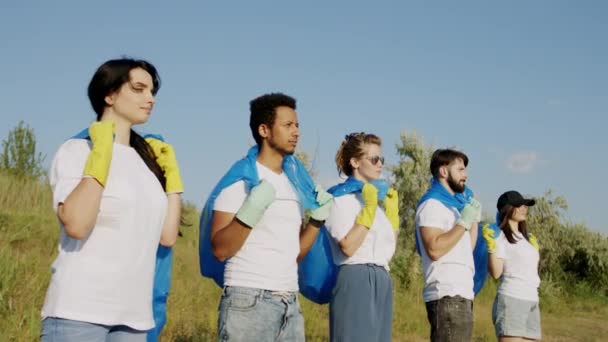 Close-up van het cameraportret van multi-etnische vrijwilligers die aan de strandkant staan en blauwe plastic tassen klaar houden om het afval op te ruimen — Stockvideo