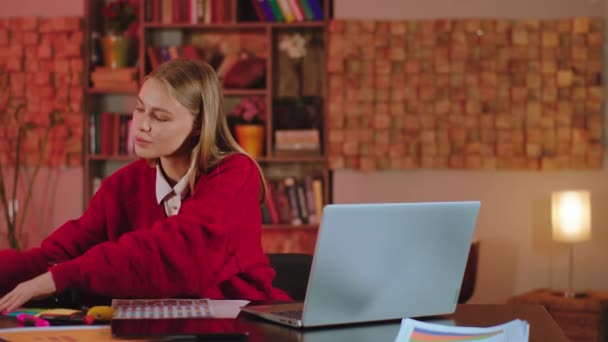 快乐聪明的女学生坐在图书馆里写一些关于她的大学项目的笔记。她笑得又可爱又大，穿着红色毛衣 — 图库视频影像
