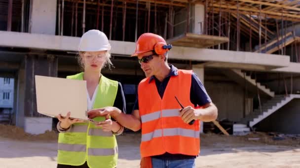 Foreman středního věku a jeho asistentka dáma kráčející před kamerou s notebookem analyzovat plán budování diskuse spolu mají na sobě bezpečnostní přilby a brýle — Stock video