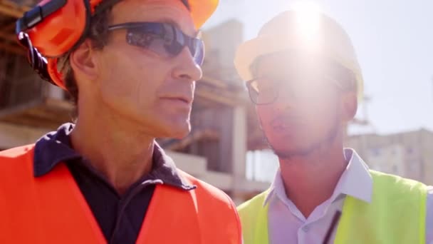 Afroamerikanischer Ingenieur und eine andere Gruppe verschiedener Spezialisten auf der Baustelle analysieren gemeinsam den Bauplan anhand der Ration, um mit anderen Spezialisten von oben zu sprechen — Stockvideo