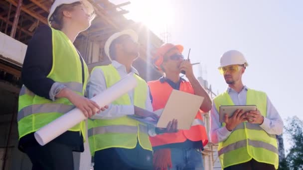 Volledige uitrusting team multi-etnische van de bouw in het midden van de bouwplaats met behulp van het rantsoen om een gesprek te hebben met andere bouwvakkers van het dak — Stockvideo