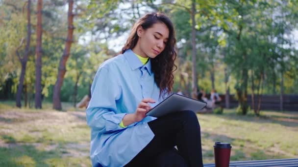 Красива азіатська леді посеред парку сідає на стілець і за допомогою електронного планшета вона вводить щось спілкування з кимось під час кави — стокове відео
