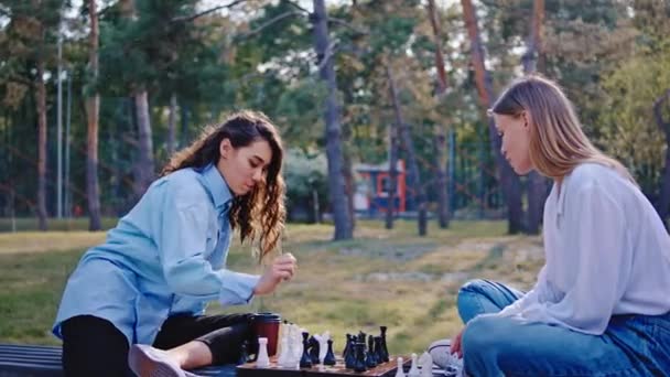 Chicas guapas en un parque moderno al aire libre jugando juntas en un juego de ajedrez muy concentrado sonriendo y sintiéndose felices — Vídeo de stock