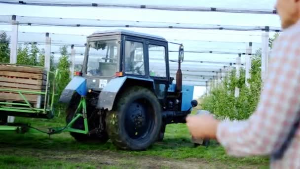 Tractor de huerto moderno y desarrollado conduciendo con una cosecha llena de manzanas a través del huerto mientras el hombre agricultor supervisa — Vídeo de stock