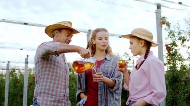 In de appelboomgaard neemt een gelukkige boer een kruik met een vers biologisch sap en giet in een aantal glazen voor twee landelijke dames — Stockvideo