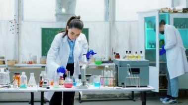 Kameranın önünde konsantre olmuş çalışan biyokimyacı kadın ve adam bir test deneyi yapıyorlar. Bir damlatıcı kullanarak bir miktar kimyasal sıvı ekliyorlar.