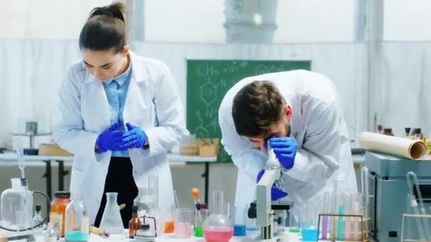 Przed kamerą dwie koleżanki naukowiec kobieta i mężczyzna w laboratorium chemicznym analizując coś przez mikroskop pracują nad czymś. Strzał na ARRI Alexa Mini. — Wideo stockowe