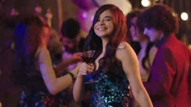 Bir şeyler partisinde dans eden kadın kameranın önünde rahat ve mutlu bir şekilde bir bardak içki tutuyor ve gülümsüyor.