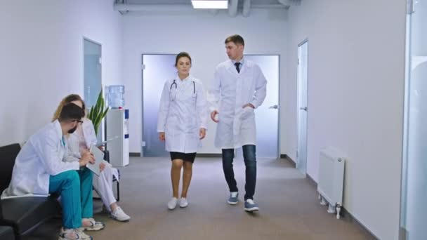 V moderní nemocniční chodbě procházející před kamerou dva lékaři žena a muž mají diskusi o některých problémech pacienta pozadí jiné nemocniční věci se posaďte na — Stock video