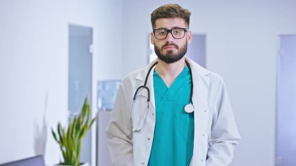 Geweldig uitziende man dokter in een modern ziekenhuis gang met een bril recht naar de camera kijkend concentreerde hij zich op zijn nek de stethoscoop — Stockvideo