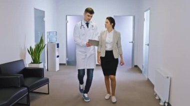 Hastane koridorunda yürüdükleri sağlık kontrolünden sonra hastane hanımı ve yetişkin doktoru kameranın önünde tartışıyorlar.