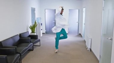 Modern hastane koridorunda kameranın önünde büyük bir gülümsemeyle dans eden heyecanlı doktor çok mutlu.