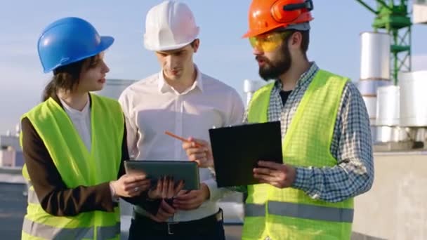 In front of the camera group of specialists in construction site they analyzing the plan of the building using digital tablet they wearing all the safety equipment — Stock Video